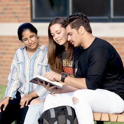 Three college students sitting on bench outside reading Bible