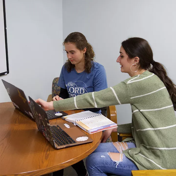 Female student tutor helping another female student using laptop computers and textbook seated at a table.